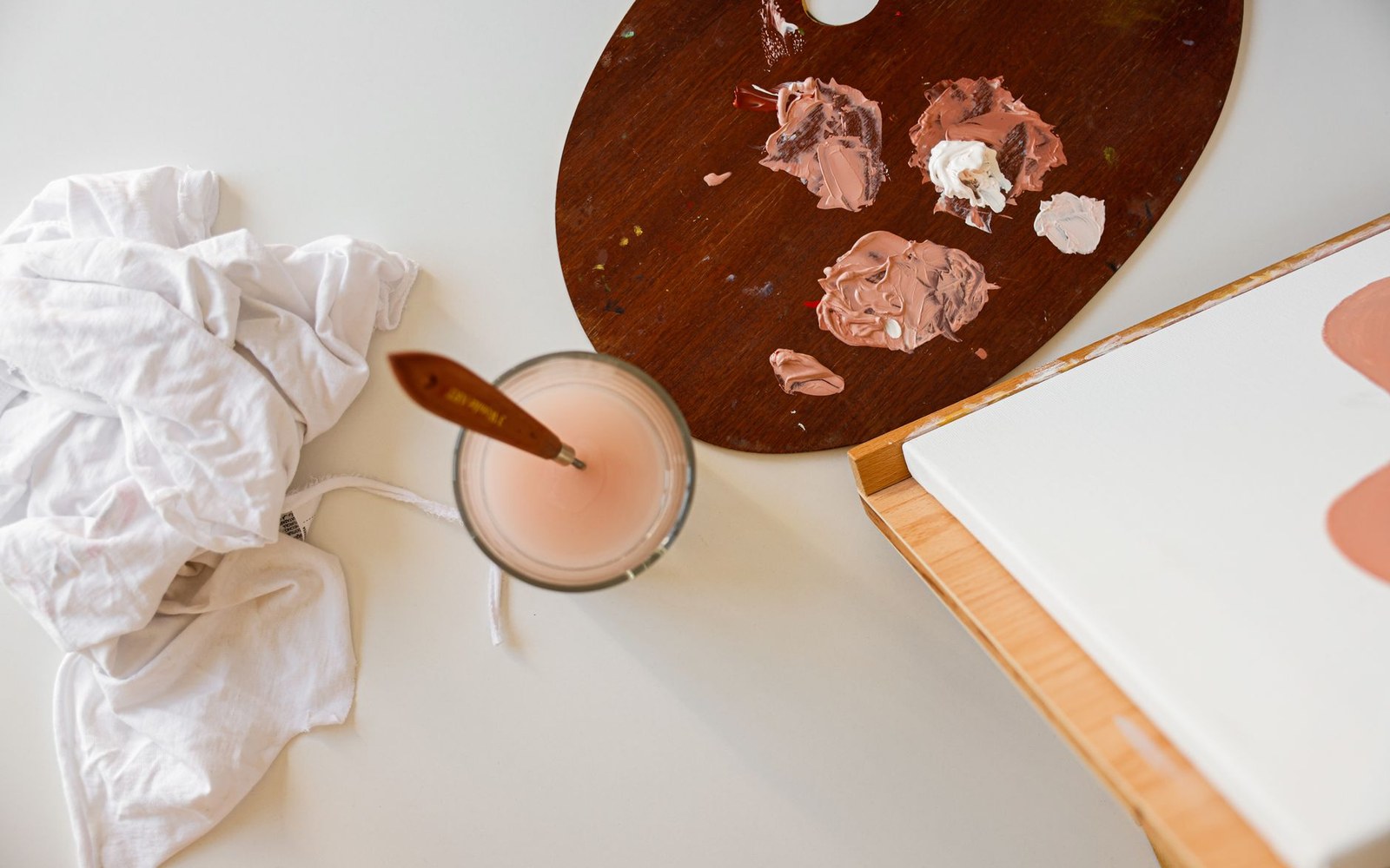 Wooden palette with pink and white paint, brush in water jar, and canvas edge