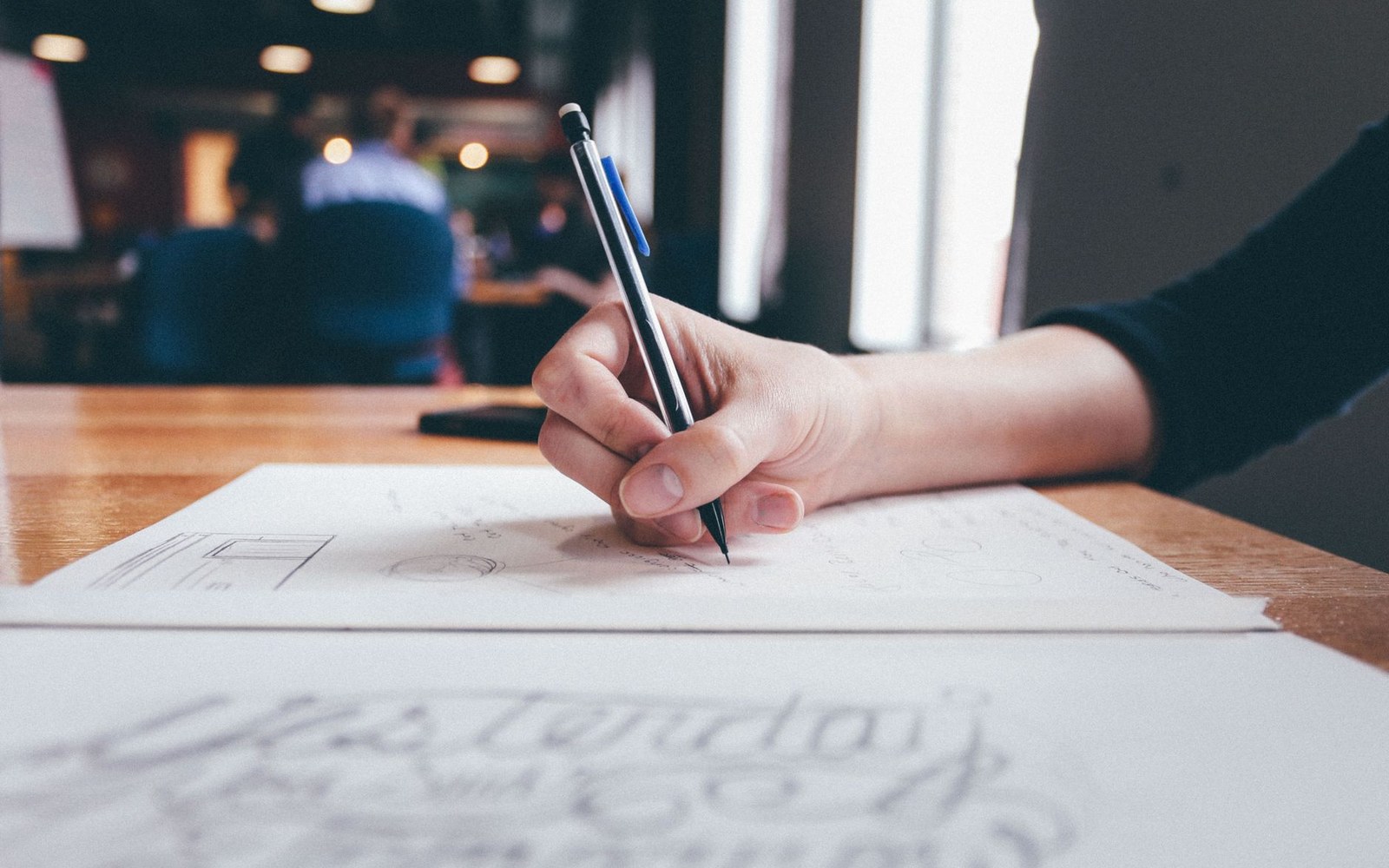 A hand sketches with a pen on paper at a wooden table in a café
