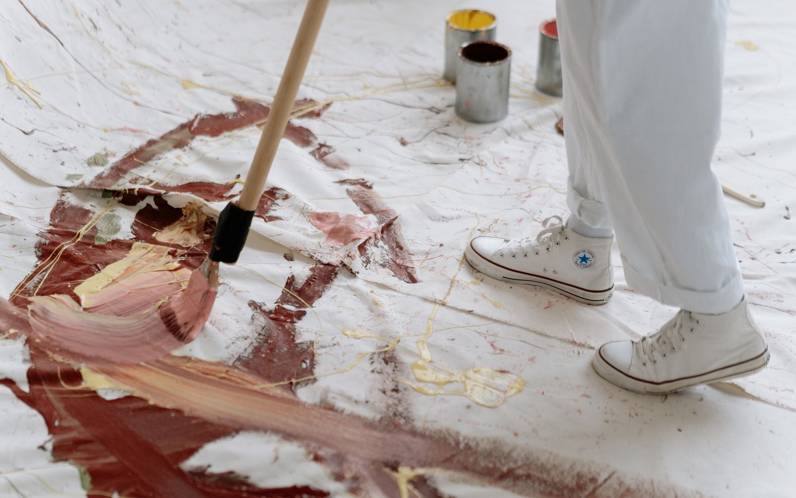 A person in white sneakers drags a large brush across paint-covered canvas on the floor