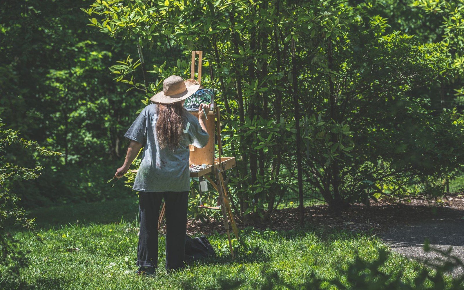 A wide-brimmed hat painter works at an easel amid dense greenery outdoors