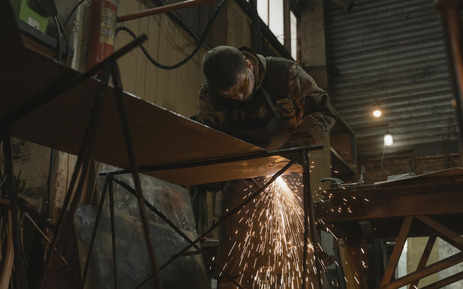 Worker in camouflage clothing grinds a metal plate, sending sparks flying in a workshop
