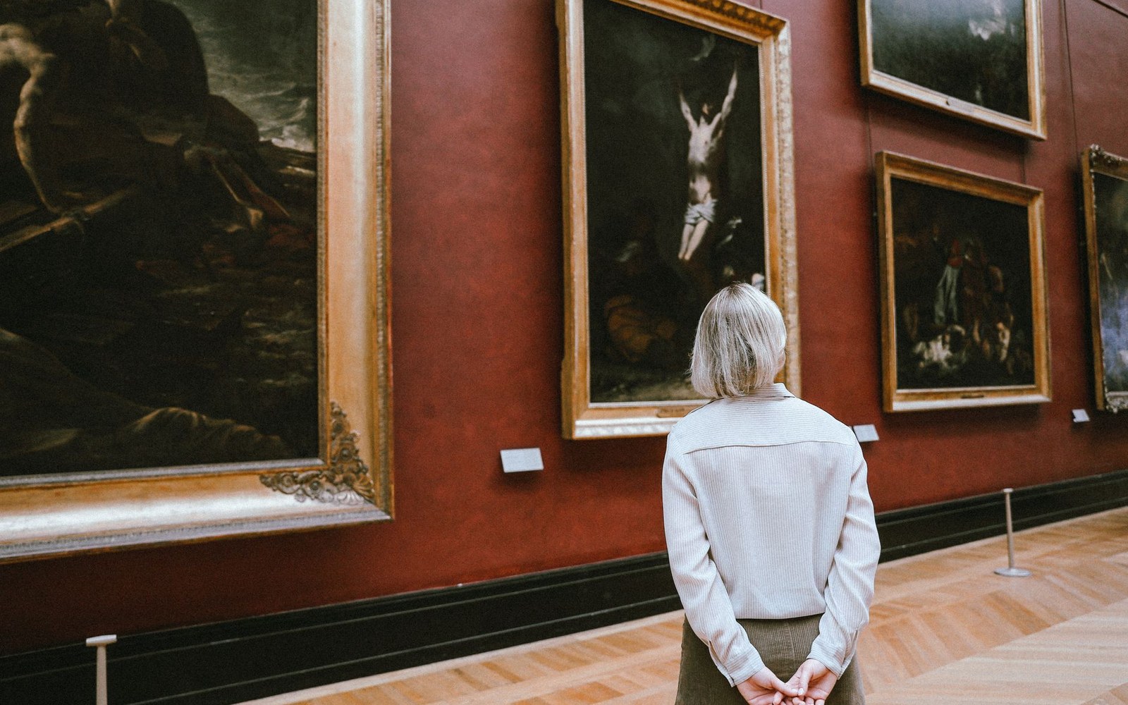 A woman studies gilt-framed paintings on a red gallery wall