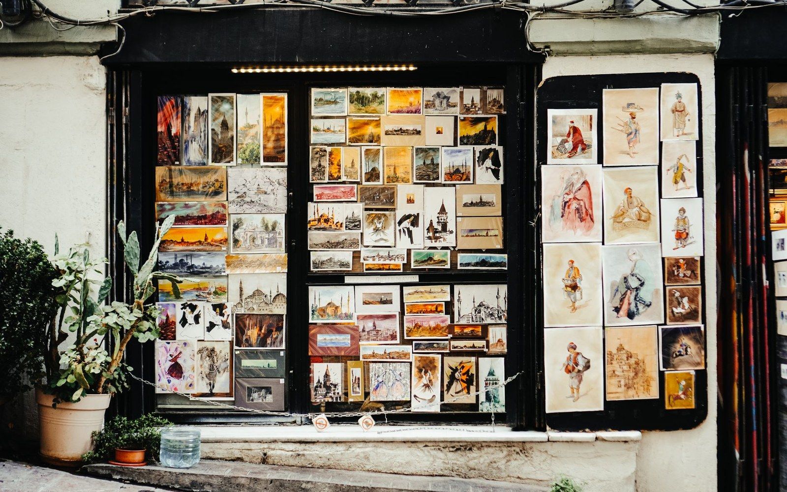 Postcards and paintings cover a shop's windows and door, with potted plants outside