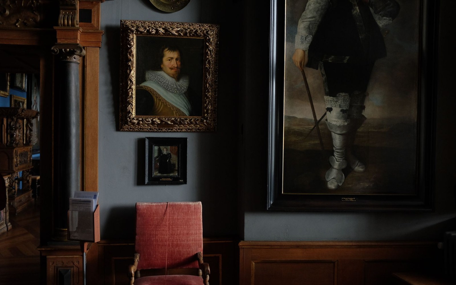 Gilt-framed portraits hang above a velvet armchair in a dimly lit gallery room