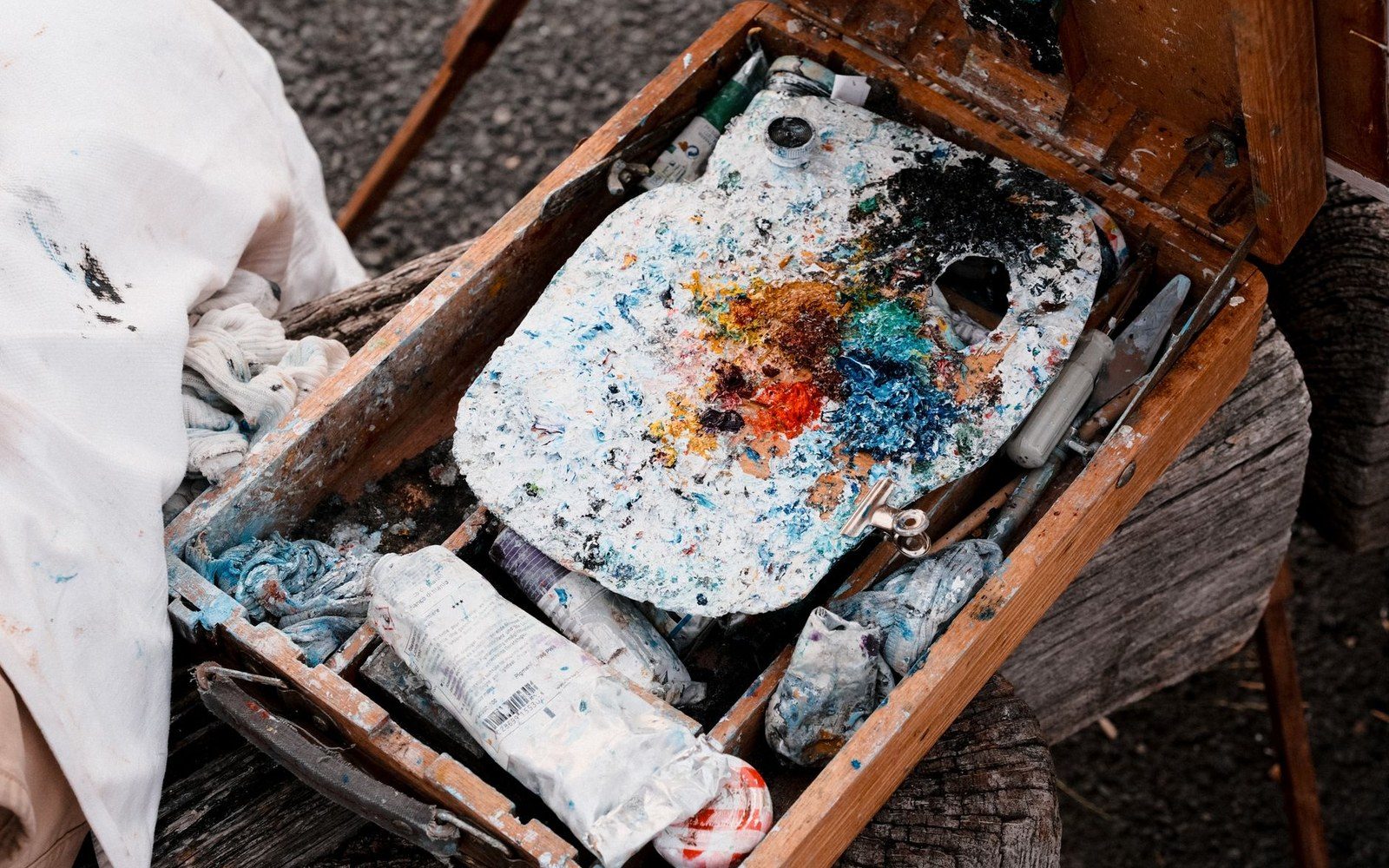 A paint-splattered palette rests inside a wooden field easel box with used paint tubes