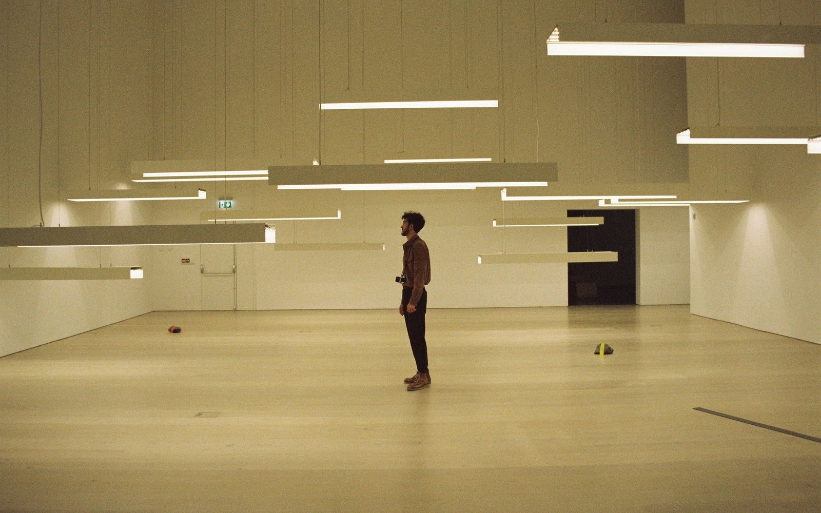 A man stands alone in an empty gallery beneath rows of suspended fluorescent light fixtures